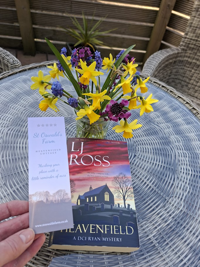 alt="the book Heavenfield on a table with bookmark and flowers behind"