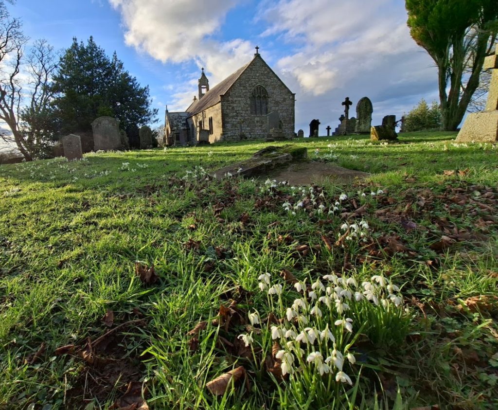 alt="Heavenfield Church and churchyard with blue skies and snowdrops around the gravestones"